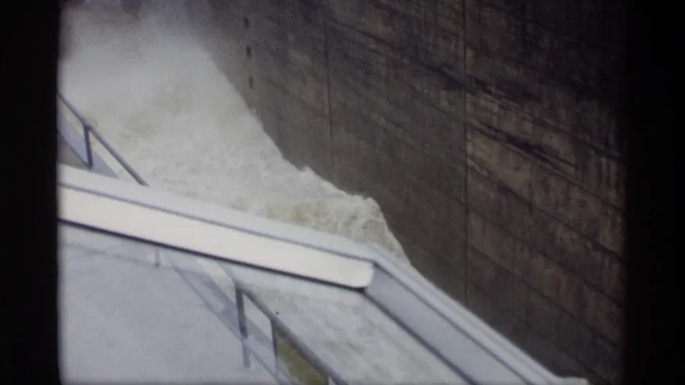 1969 Savage, Minnesota 8mm frame showing rushing water near a concrete embankment and railing beneath a dark bridge wall.