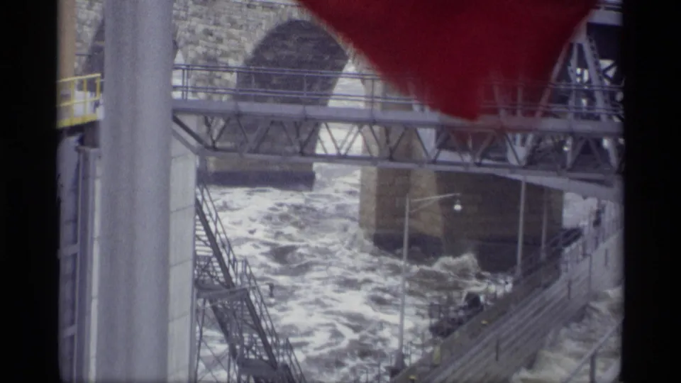 1969 Savage, Minnesota 8mm frame with foaming river water, a stone arch bridge, metal truss span, railings, and a concrete support.