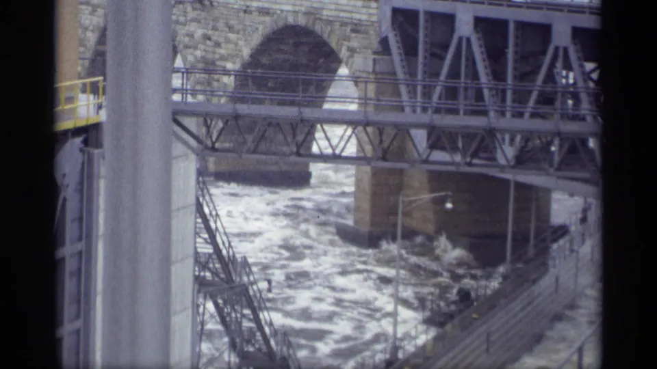 1969 Savage, Minnesota 8mm frame showing stairs, railings, trusswork, a stone arch, and turbulent water in a spillway channel.
