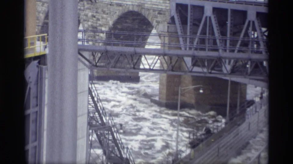 1969 Savage, Minnesota 8mm frame of fast river water under a metal truss walkway with railings, a stone arch, and concrete supports.