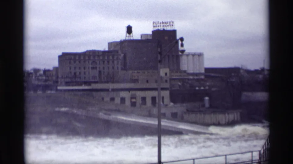 1969 home-movie frame of a Savage, Minnesota industrial riverfront with a Pillsbury's sign above rushing water.