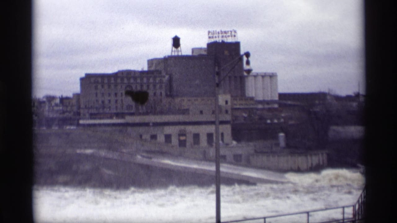 River water below industrial buildings with a grain elevator and signage in the distance.