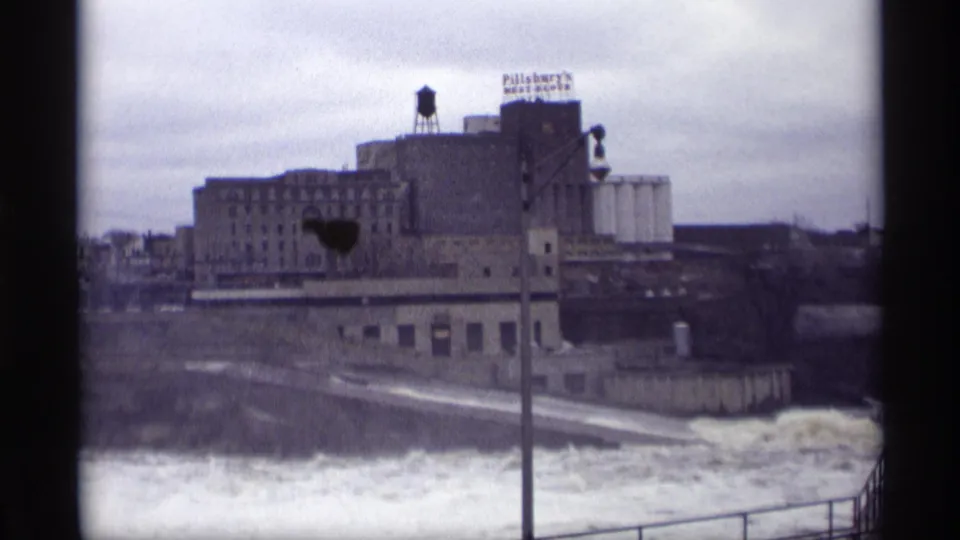 1969 8mm view in Savage, Minnesota, showing turbulent river water below large industrial and grain elevator buildings.