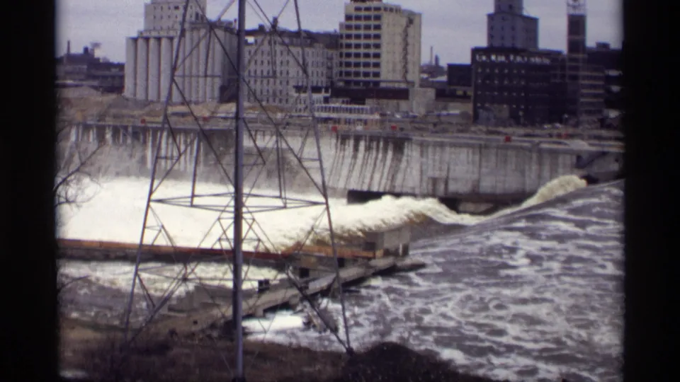 Savage, Minnesota riverfront view from 1969 with dam outflow, a metal transmission tower, grain elevators, and industrial buildings.