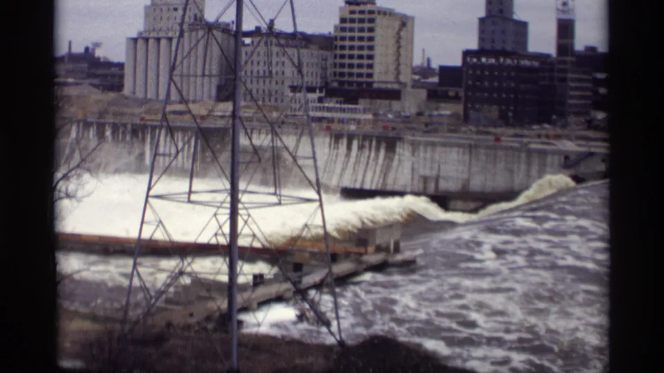 1969 8mm frame showing foaming dam water in Savage, Minnesota, with a tower in the foreground and industrial buildings behind.