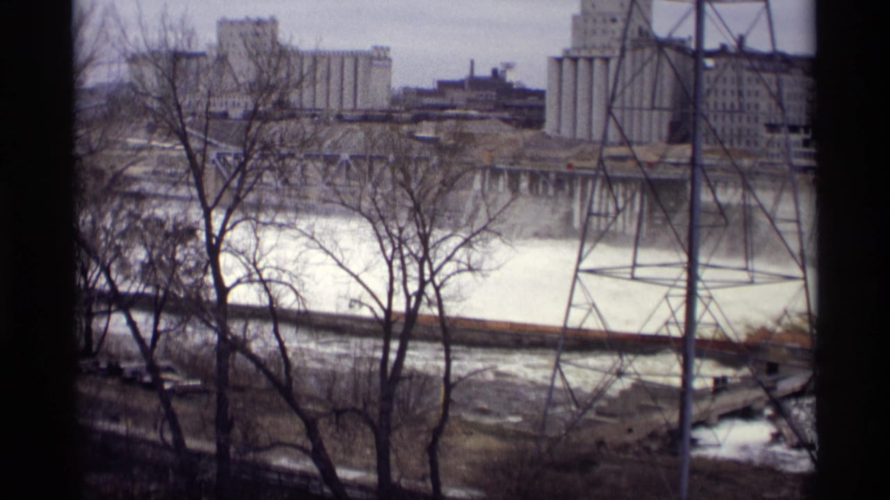 1969 8mm view in Savage, Minnesota of fast river water framed by bare trees, with industrial structures and a power tower across the river.