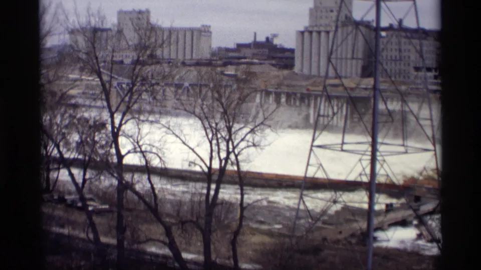 1969 8mm view in Savage, Minnesota of fast river water framed by bare trees, with industrial structures and a power tower across the river.