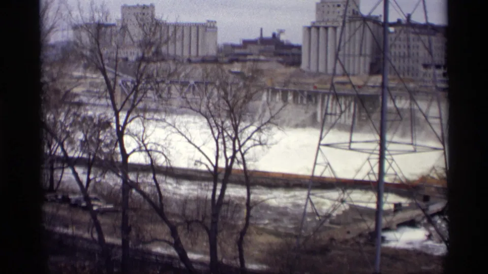 Savage, Minnesota riverfront in 1969, showing rushing water, leafless trees, industrial buildings, and a tall steel transmission tower.