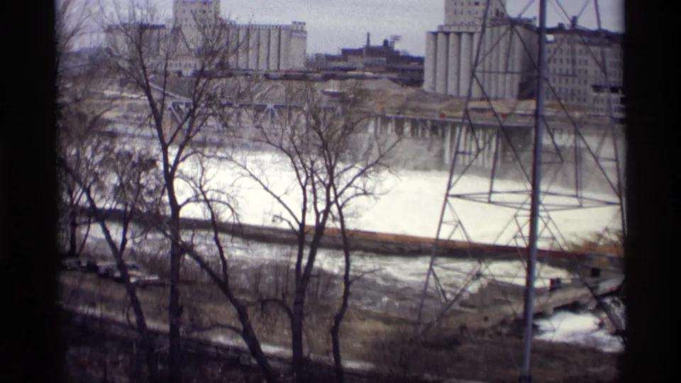 1969 8mm frame of turbulent river water in Savage, Minnesota, with bare foreground trees and industrial structures across the river.