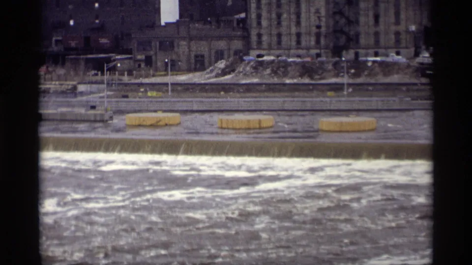 Savage, Minnesota river scene from 1969 showing whitewater over a low barrier, shoreline posts, snow or pale debris, and industrial buildings beyond.