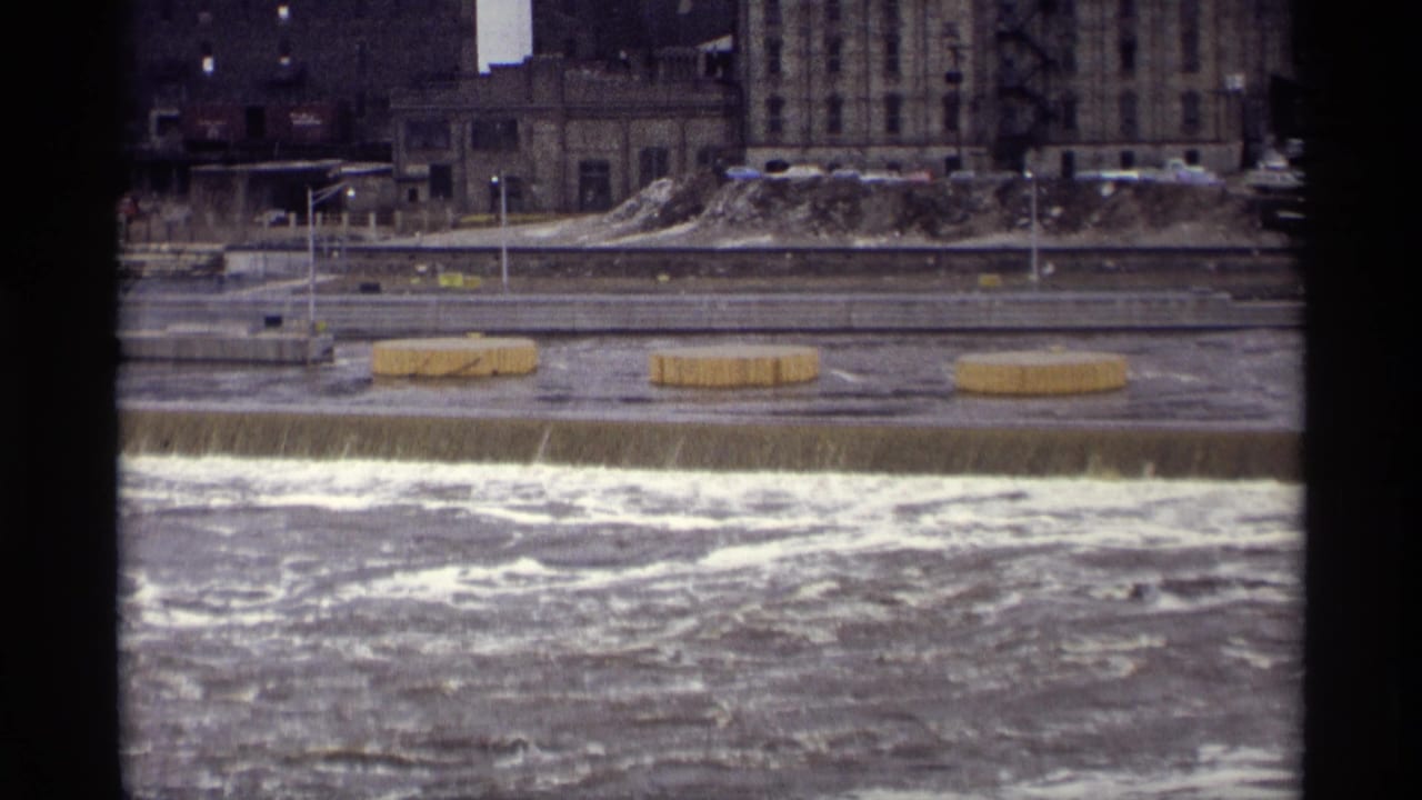 Foaming river water flows over a low dam with buildings across the river.