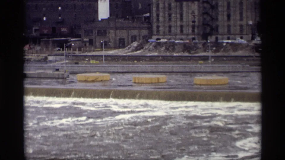 1969 Savage, Minnesota 8mm frame of white water below a concrete spillway, with several cylindrical structures in the current.
