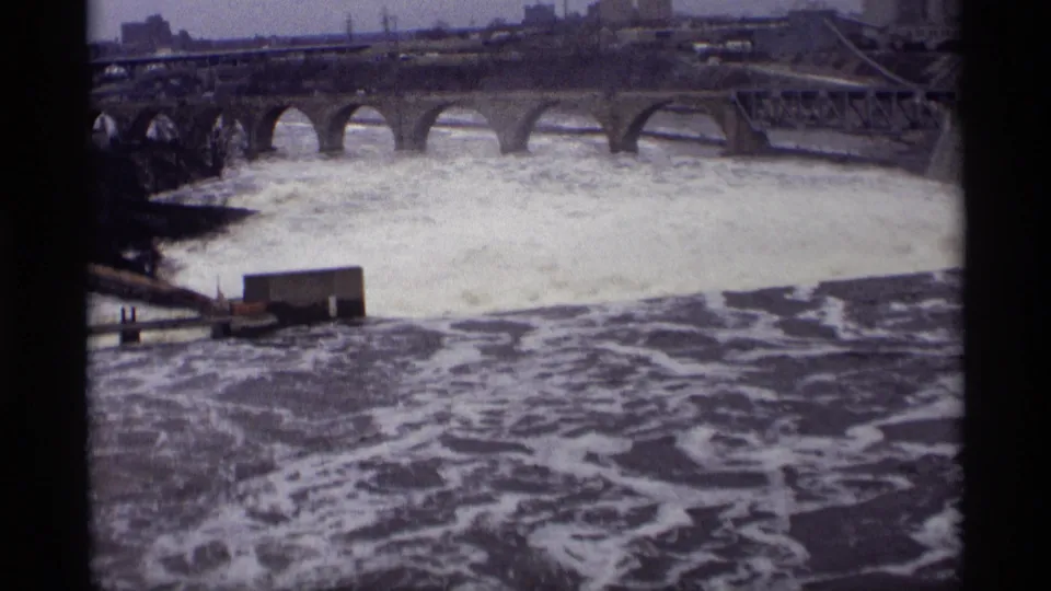 1969 Savage, Minnesota 8mm frame showing a broad foaming river and a multi-arched bridge beyond the dam area.