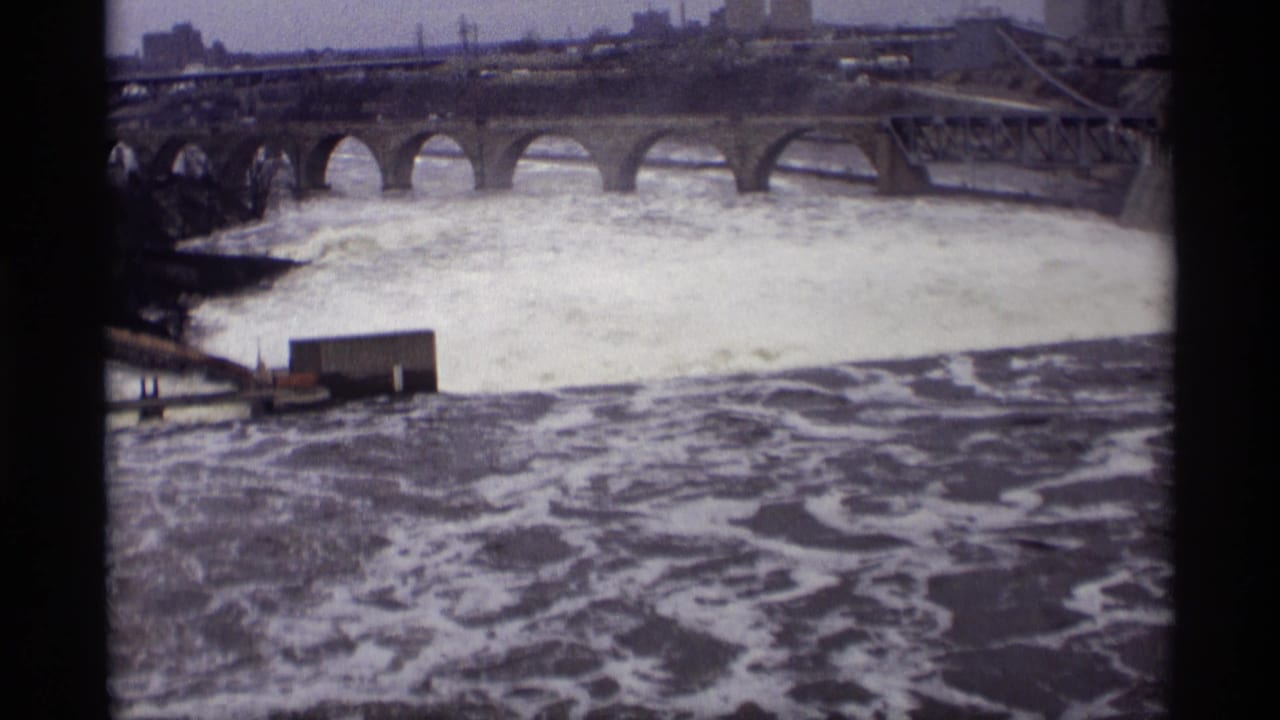 1969 8mm view in Savage, Minnesota, showing rough river water below a stone arch bridge with industrial structures beyond.
