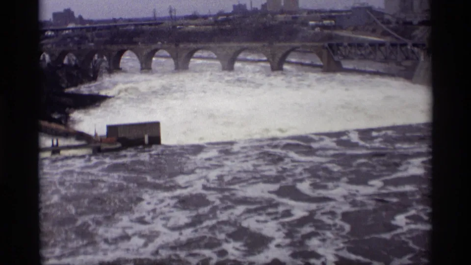 1969 8mm view in Savage, Minnesota, showing rough river water below a stone arch bridge with industrial structures beyond.