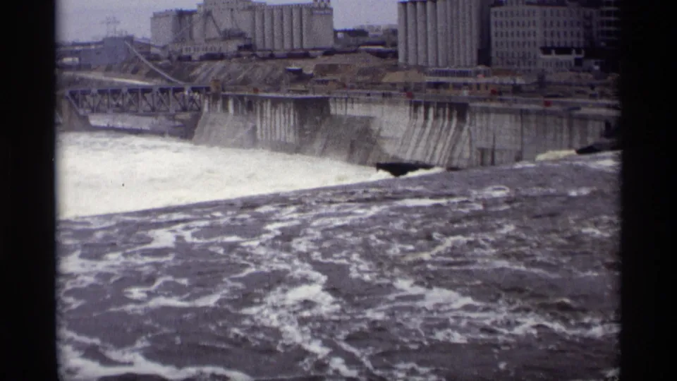 Savage, Minnesota riverfront view showing a concrete dam spillway, churning water, grain elevators, and industrial buildings in 1969 footage.