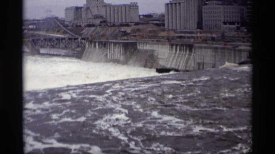 Late-1960s 8mm frame of the Savage, Minnesota dam area, with heavy whitewater in front of grain elevators and industrial buildings.