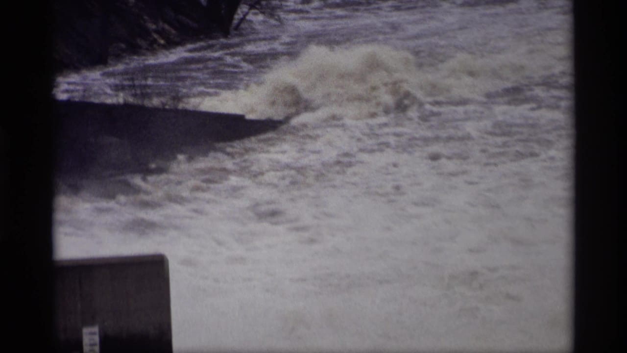 Large wave rolls over churning water near a concrete riverbank structure.