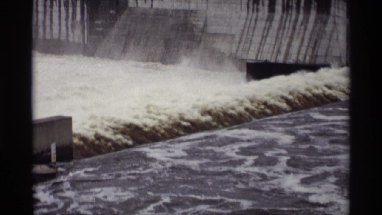 Foaming water surges over a concrete spillway with dark outlet openings above.
