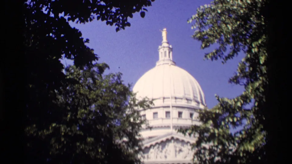 1969 Wisconsin home-movie frame of a white domed landmark seen through trees under clear blue sky.