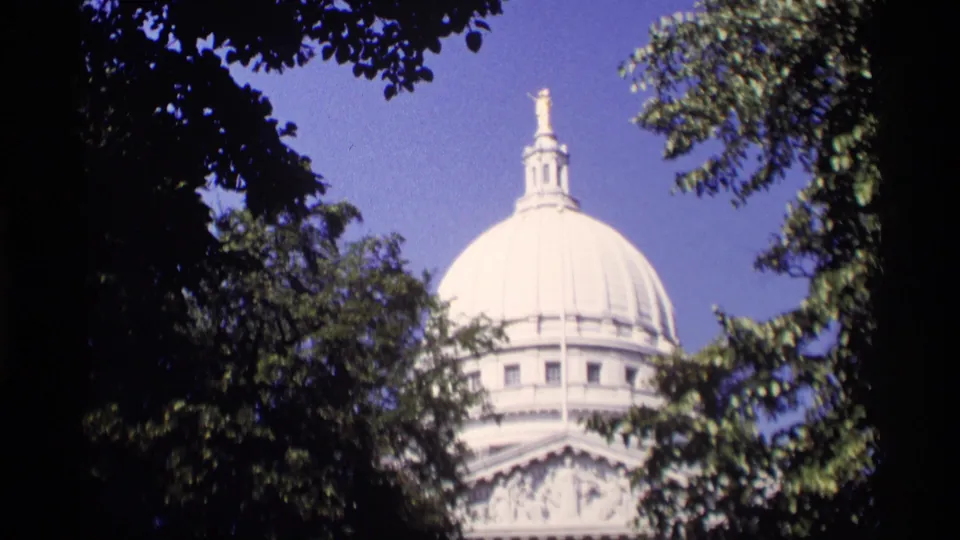 1969 8mm view of the Wisconsin State Capitol dome partly framed by leafy trees and clear blue sky.