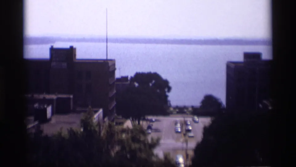 1969 Wisconsin city overlook with a street, cars, brick structures, trees, and a broad body of water beyond.