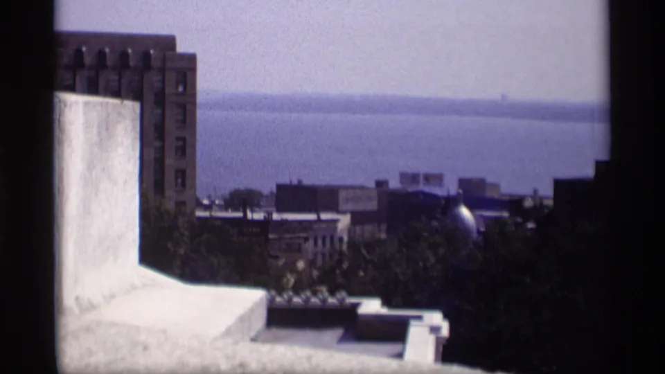 1969 Wisconsin rooftop panorama with a pale ledge, brick building, trees, low roofs, and open water beyond.