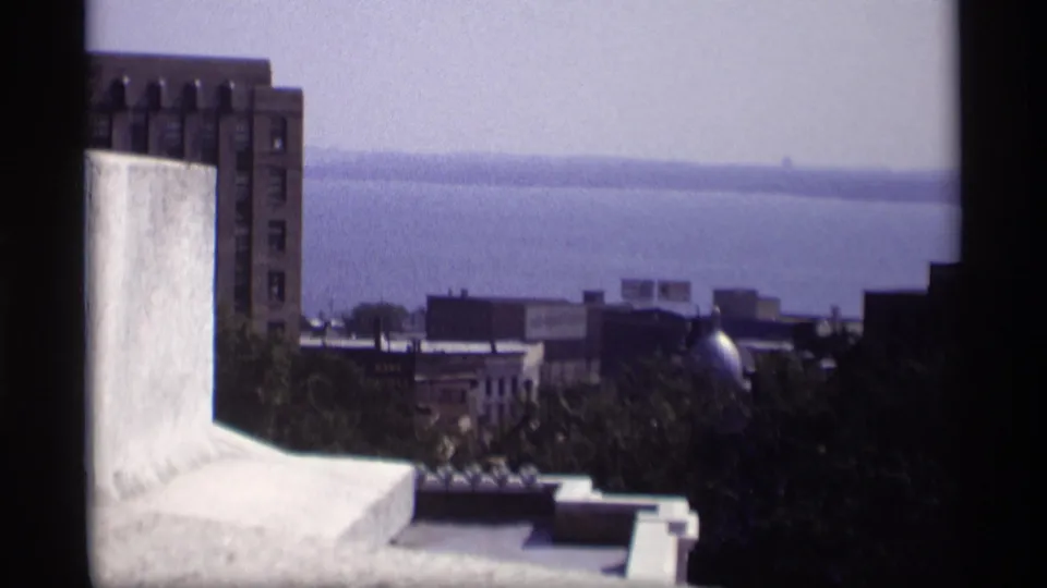 1969 Wisconsin elevated waterfront view with rooftop architecture, brick facades, trees, and distant waterline.