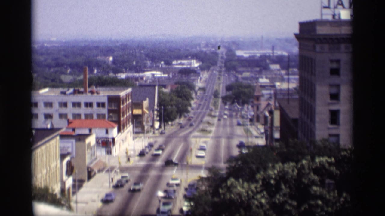 High-angle view down a long city street with cars, commercial buildings, trees, and rooftop signage.