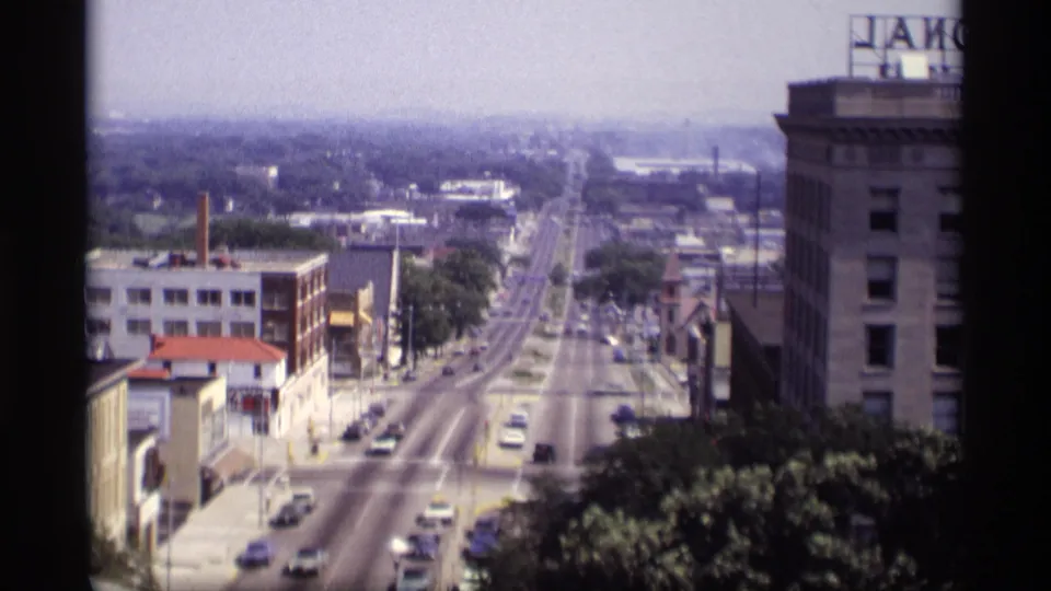 1969 Wisconsin home-movie frame of a long straight urban street with cars, sidewalks, trees, older buildings, and visible rooftop signage.
