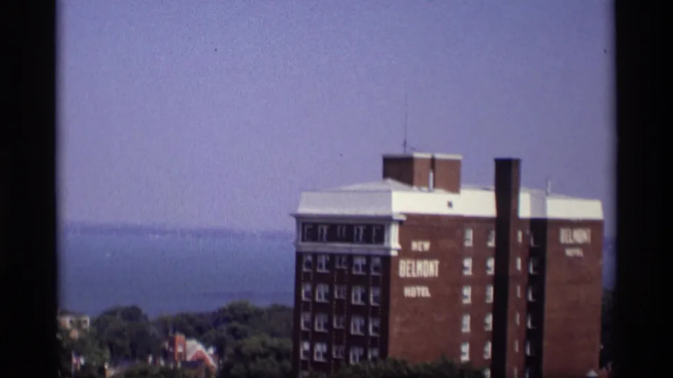 1969 Wisconsin frame showing the red brick New Belmont Hotel building near treetops, with distant water and a hazy sky behind it.