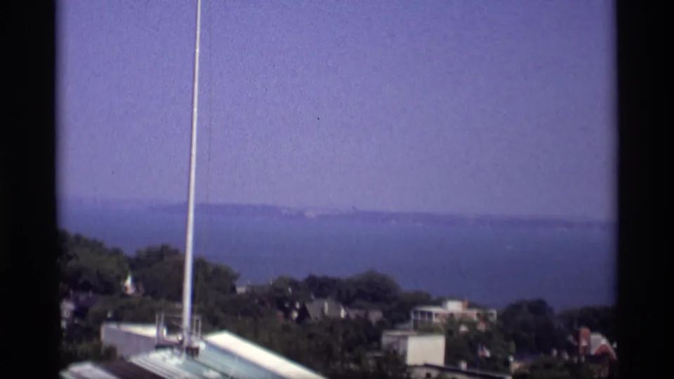 1969 Wisconsin 8mm frame continuing the pan across a flagpole, foreground buildings, trees, and a distant blue shoreline.
