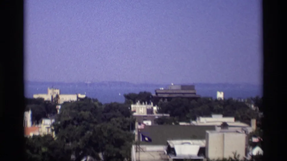 1969 Wisconsin 8mm frame showing rooftops and trees in the foreground with a broad hazy waterfront and shoreline in the distance.