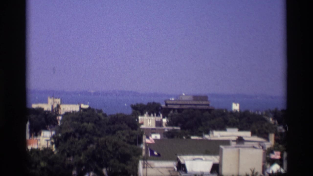 1969 Wisconsin frame with low-rise buildings and dark trees in the foreground, with water and haze beyond.