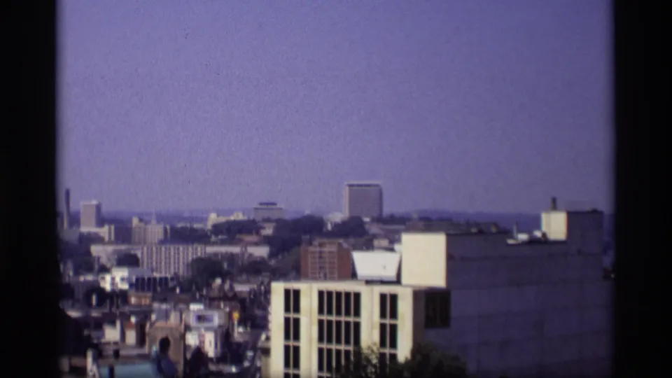 1969 Wisconsin city frame showing a higher-angle view across rooftops, trees, and taller downtown buildings in haze.