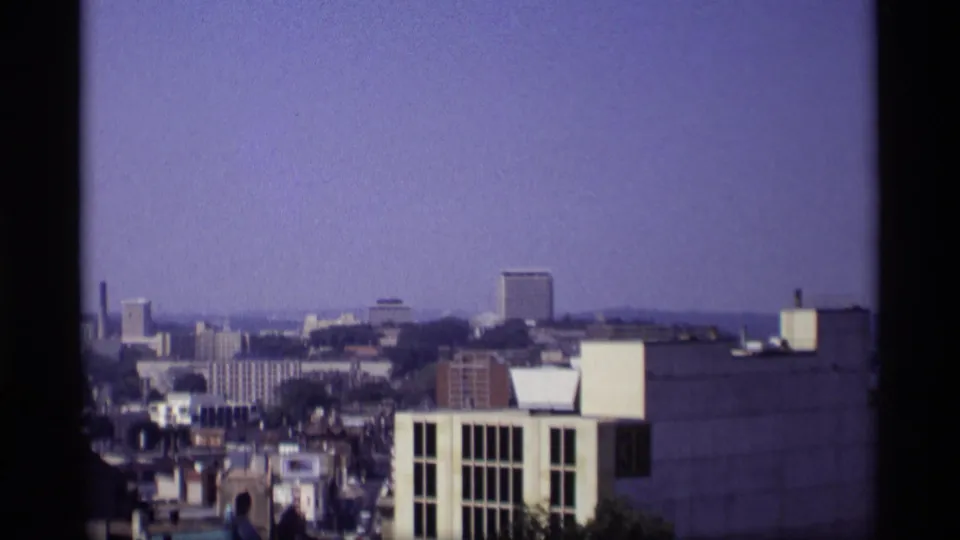 Elevated 1969 Wisconsin city view with commercial blocks, low rooftops, and dark 8mm frame edges.