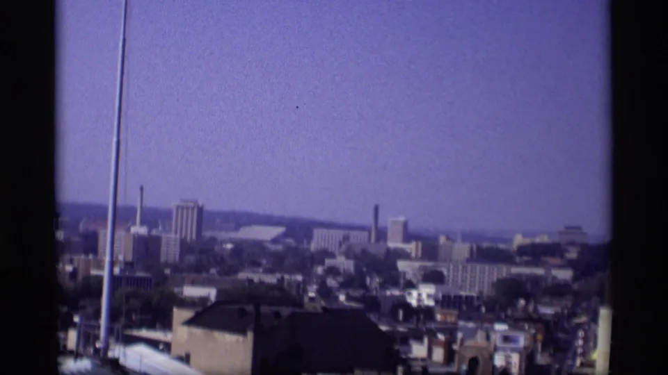 Late-1960s Wisconsin city panorama with a foreground roof, vertical pole, smokestack, and distant urban blocks.