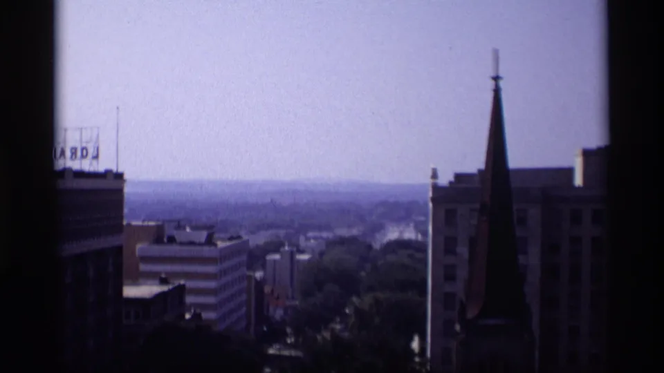 1969 Wisconsin home-movie view across a downtown skyline with a church steeple, rooftop sign, and distant tree-lined horizon.