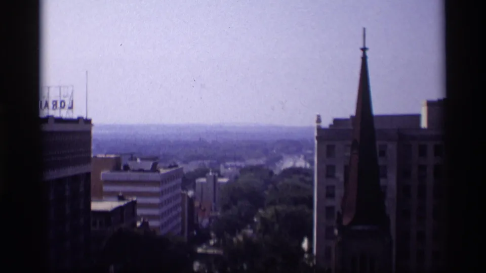 1969 Wisconsin elevated cityscape framed by a dark window edge, showing a church steeple, rooftop sign, and distant horizon.