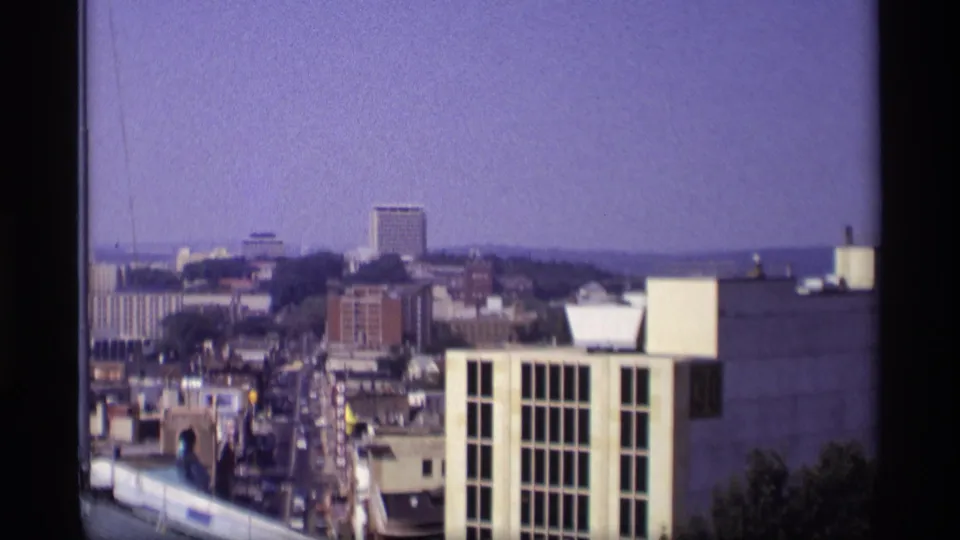1969 Wisconsin city view from above, looking along downtown buildings toward a taller block and distant wooded horizon.