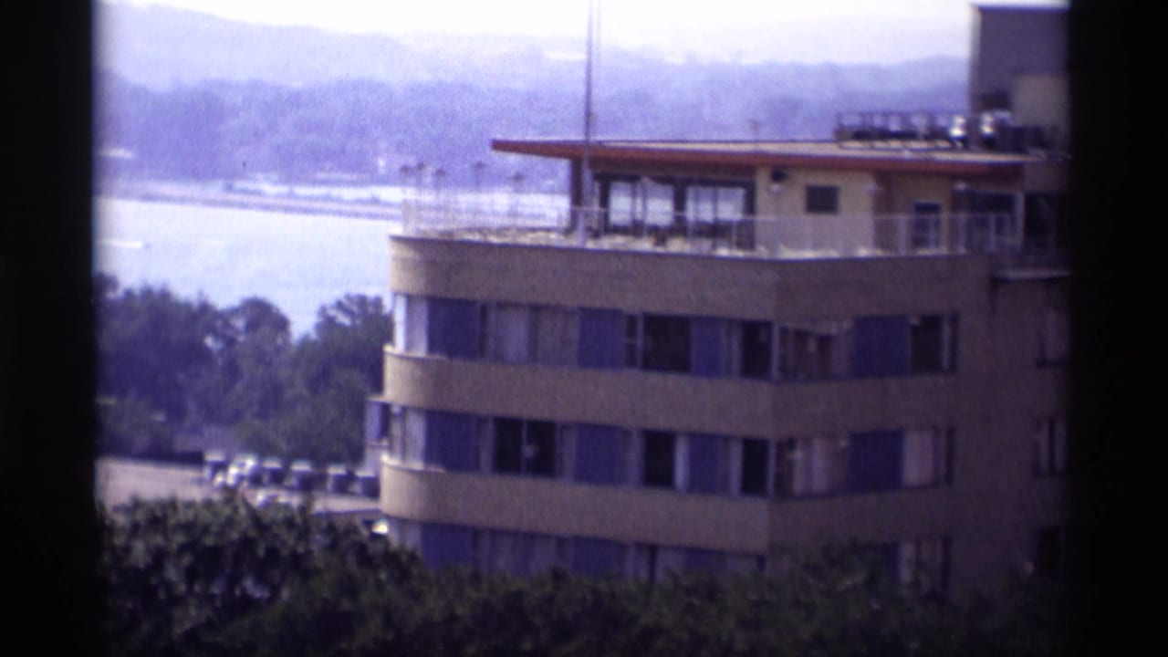 Curved multi-story building beside a river with trees and hills in the background.