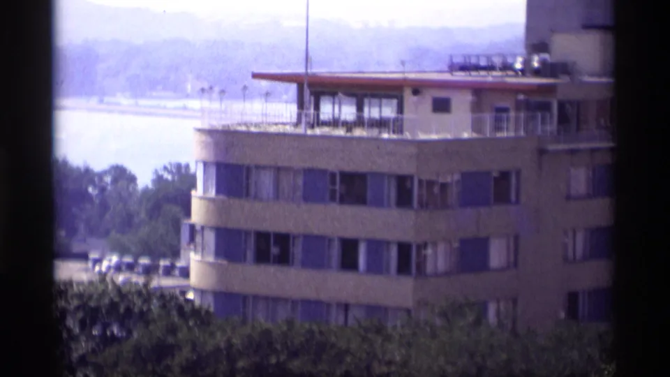Closer 1969 8mm view of a curved Wisconsin riverfront building, with rows of windows, a rooftop area, trees, and water behind it.