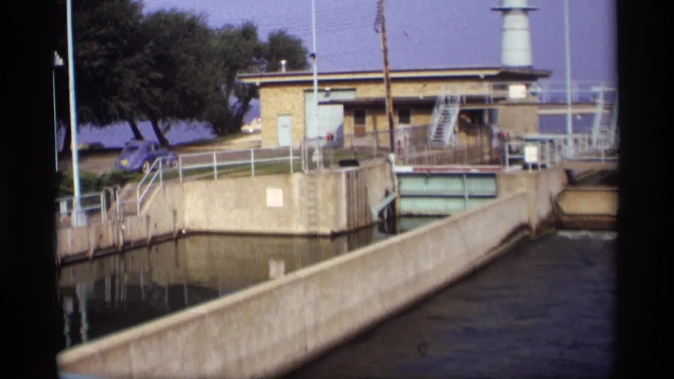 1969 Wisconsin 8mm frame showing a concrete water channel, brick control structure, metal railings, white tower, trees, and a blue parked car.