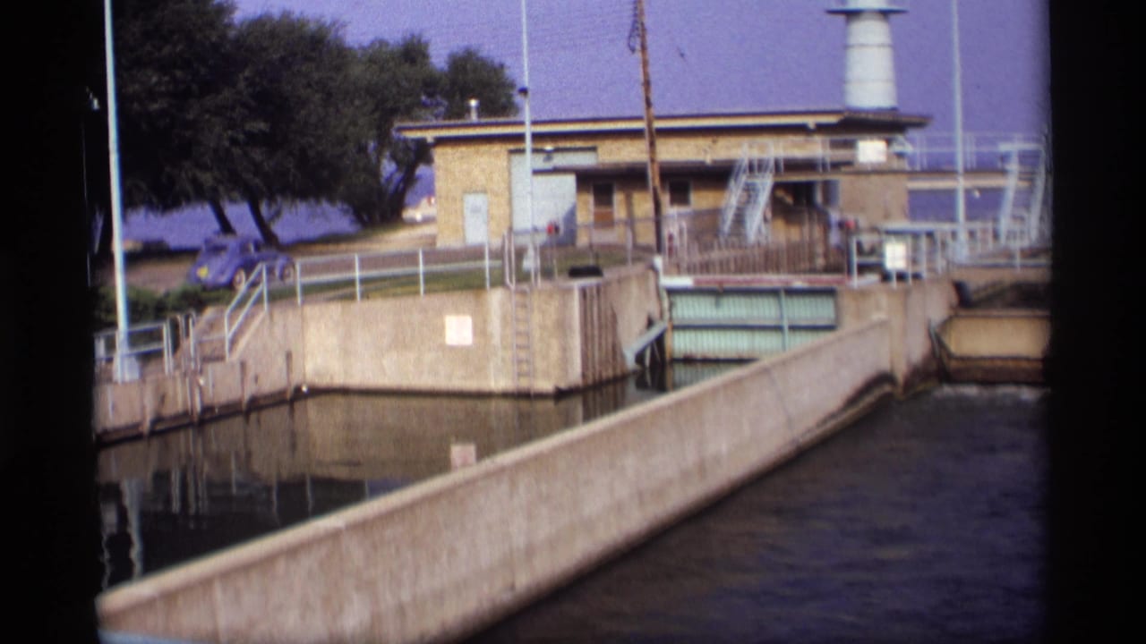 1969 Wisconsin 8mm frame of a sluice channel and industrial control station, with railings, stairs, a white tower, trees, and a blue car nearby.