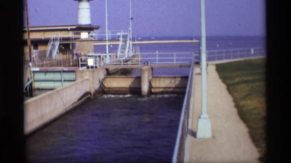 1969 Wisconsin 8mm view down a concrete canal toward a sluice gate, brick utility building, metal railings, lamppost, walkway, and open water beyond.