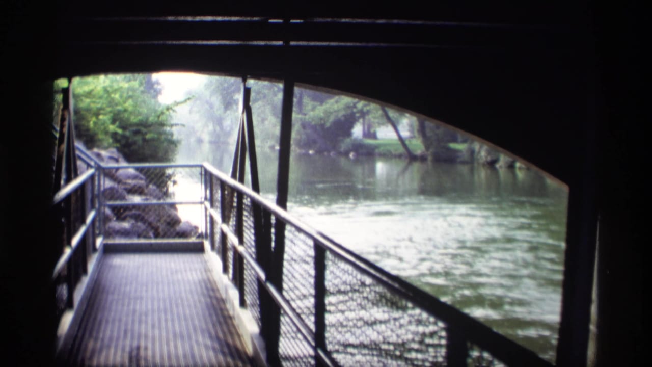 Dark bridge framing a metal walkway beside a calm river and trees.