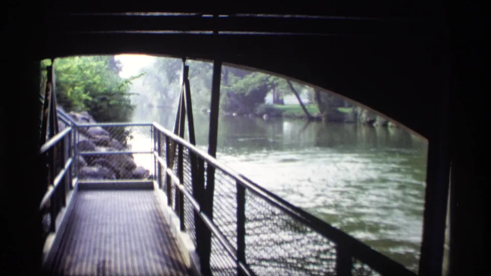 1969 Wisconsin 8mm view from beneath a bridge, with a grated walkway, railings, river water, rocks, and green trees.