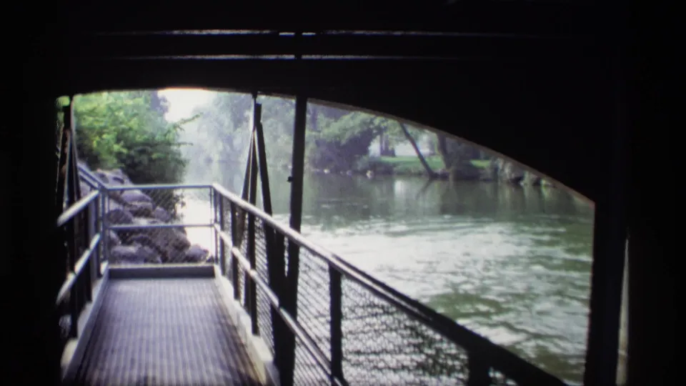 1969 Wisconsin 8mm frame looking along a riverside metal walkway from under a bridge toward water, trees, and rocks.