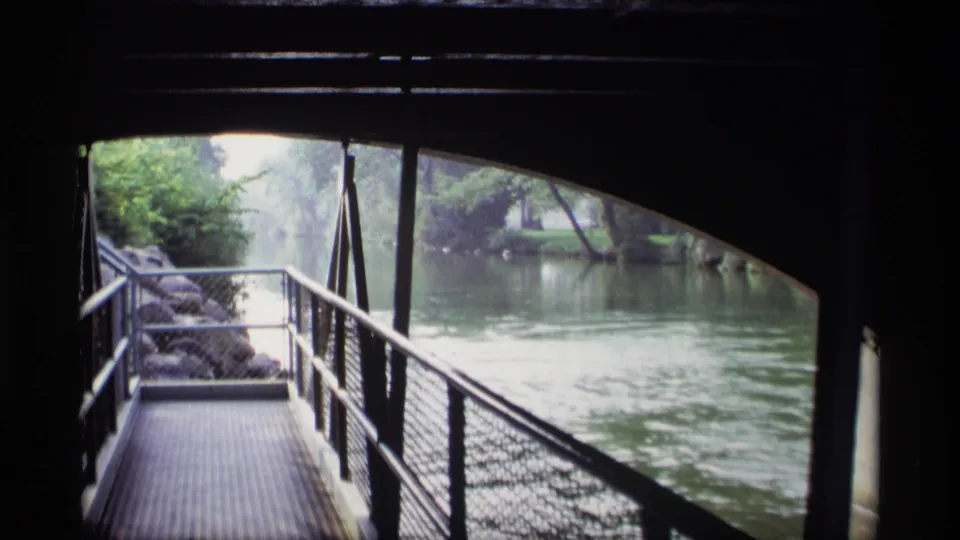 1969 Wisconsin 8mm view of a shaded bridge walkway beside a river, with railings, rocks, trees, and soft reflected light.