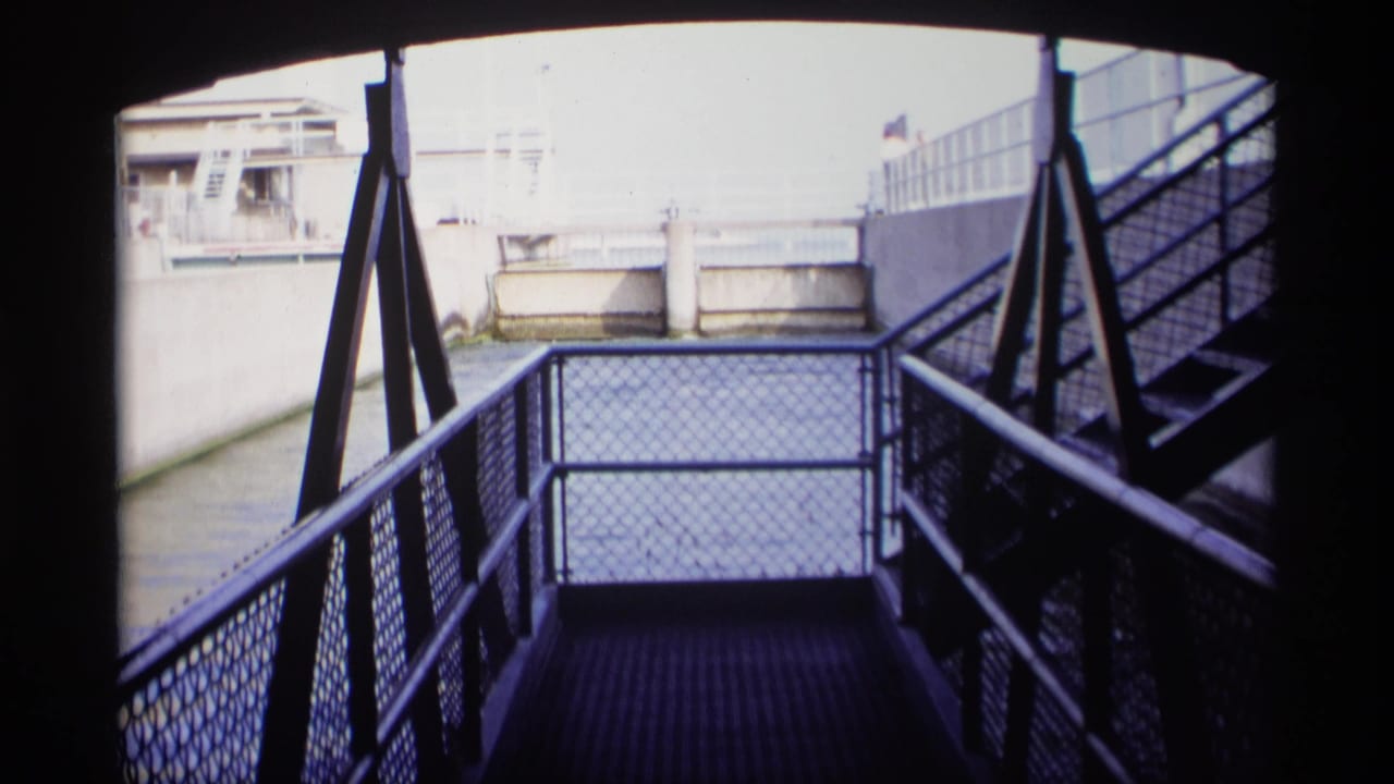 1969 Wisconsin 8mm view from a shadowed metal stair landing toward a concrete canal and pale waterfront structures.